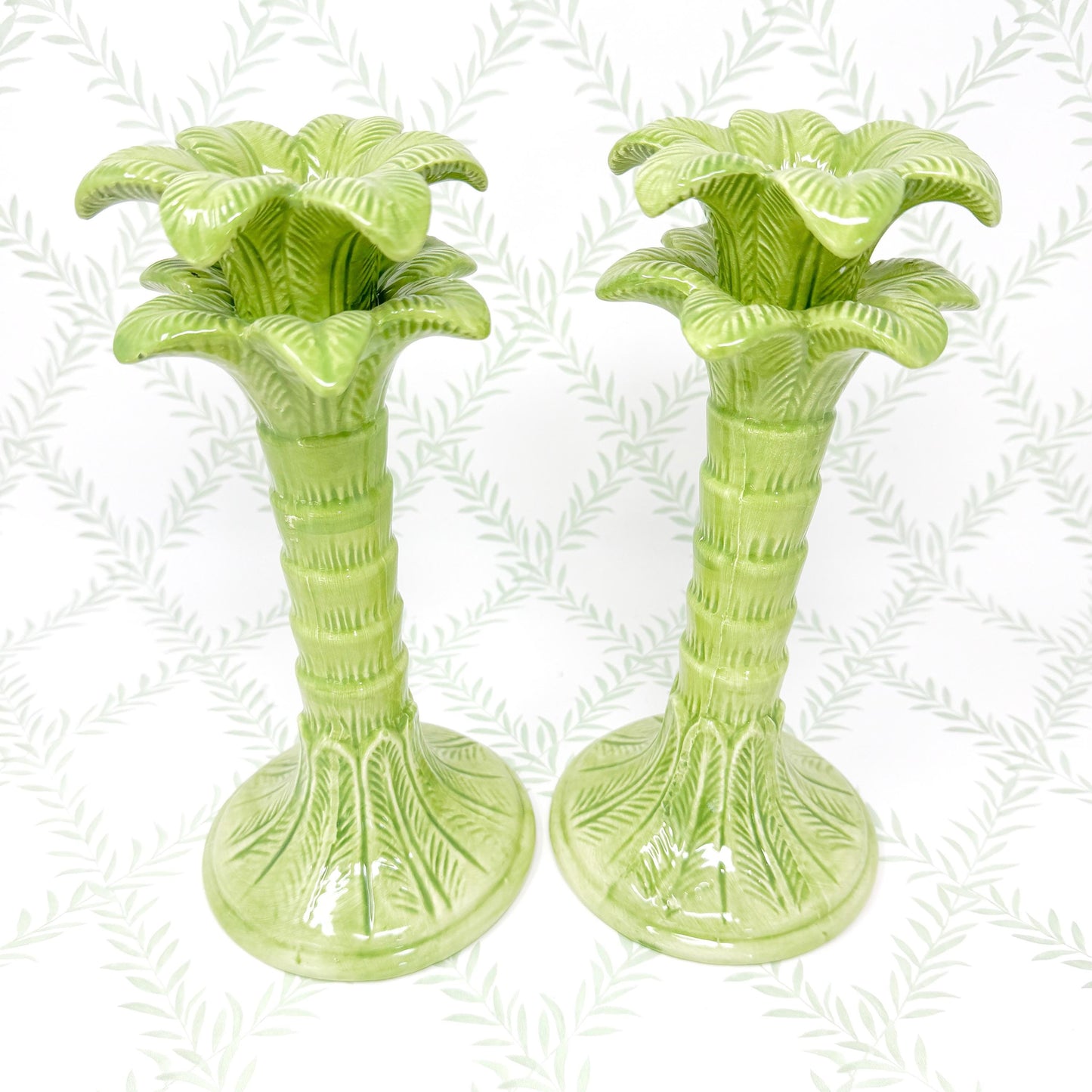 Two green ceramic palm tree candle holders. The candle holders are detailed with textured leaves and a grooved trunk. The candle holders are on a white background.