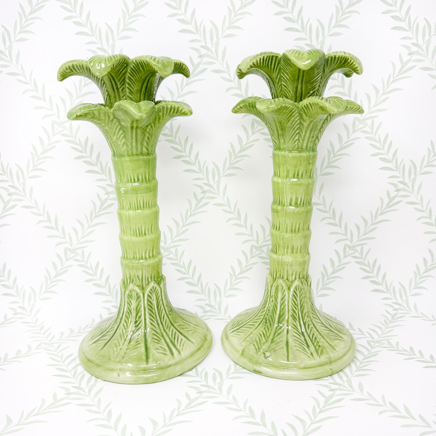 A pair of green ceramic palm tree candle holders. The candle holders are tall and slender with a wide base. The palm trees are detailed with leaves and a textured trunk.