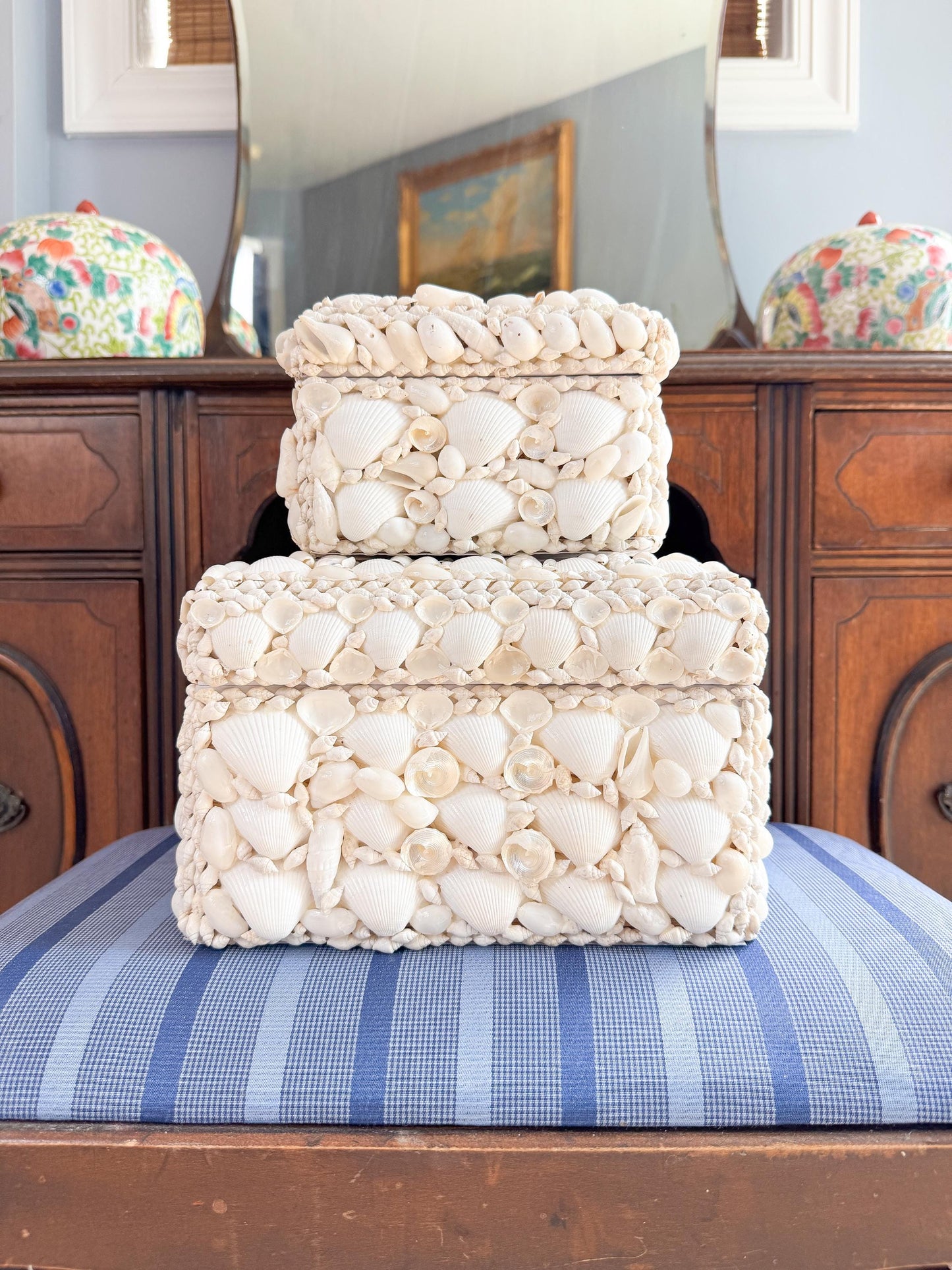 Two white square boxes made of seashells. The boxes are stacked on top of each other. The boxes are on a blue and white striped surface.