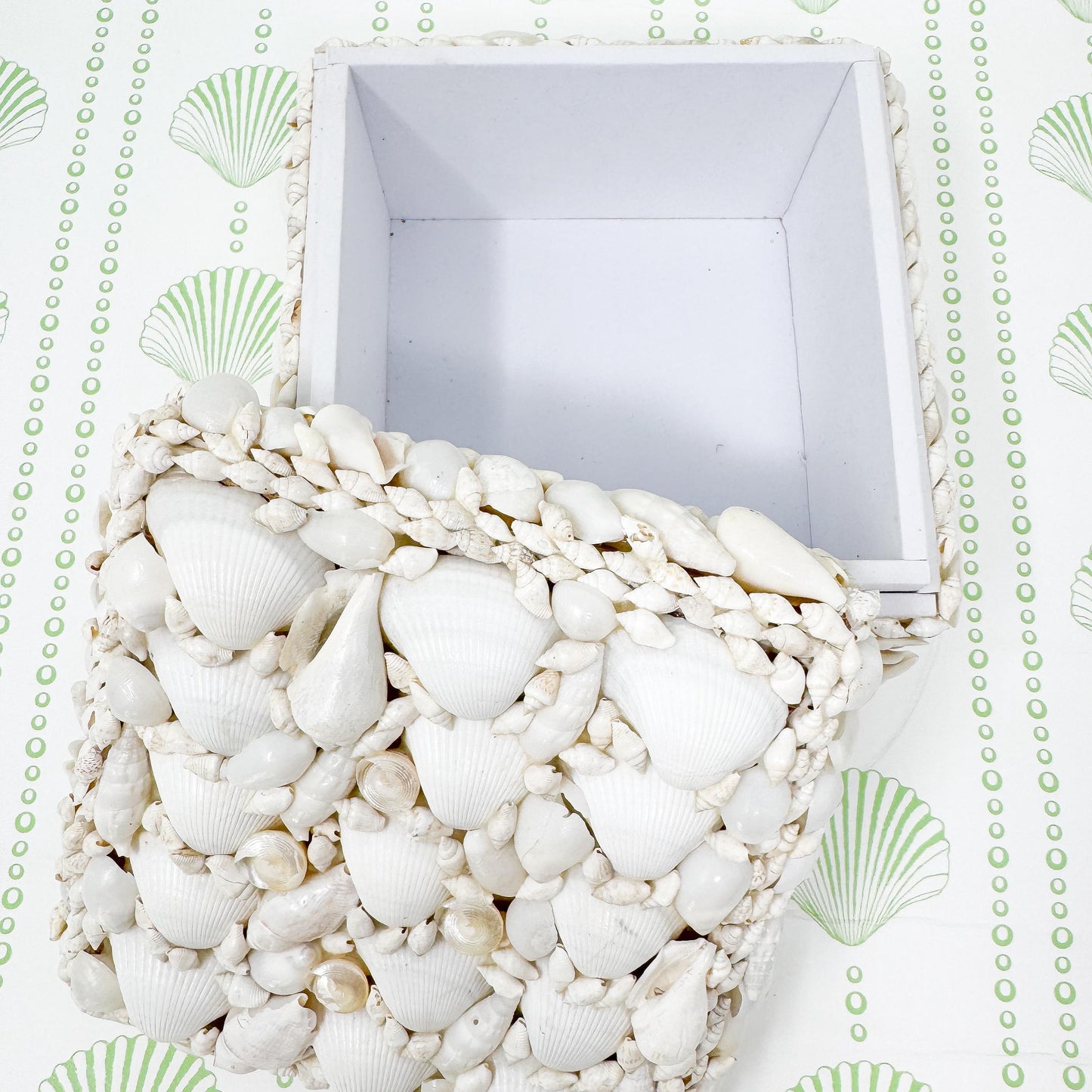 White square box with a lid made of seashells. The lid is made of a variety of seashells of different sizes and shapes. The box is open and the inside is empty and white in color.