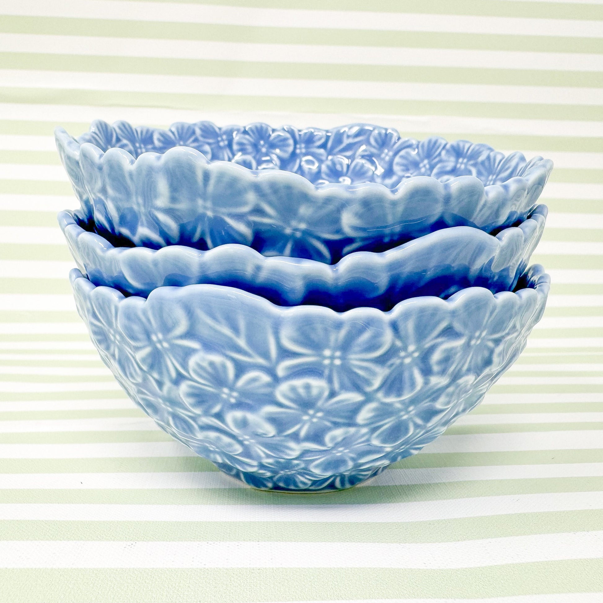 Three stackable light blue ceramic bowls with embossed floral designs.  Each bowl features a scalloped edge and a repeating pattern of small, raised flowers. The bowls are displayed against a pale green and white striped background. These decorative bowls are perfect for serving snacks, salads, or desserts.