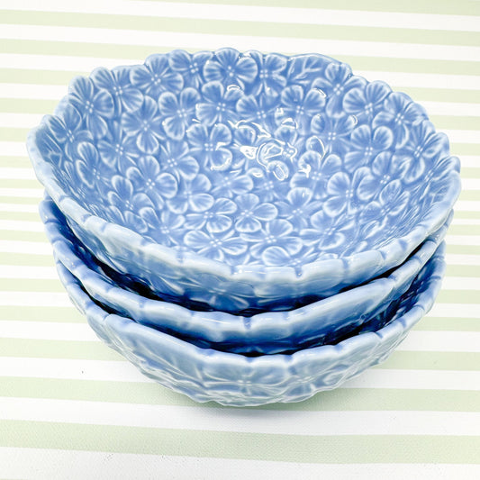 Stack of three light blue ceramic bowls. Each bowl features an embossed floral pattern of small, light blue hydrangea flowers covering the entire surface. The bowls are shallow and have a slightly scalloped edge.  The design is consistent across all three bowls, creating a cohesive set. These decorative bowls are perfect for serving snacks, salads, or as decorative accents.