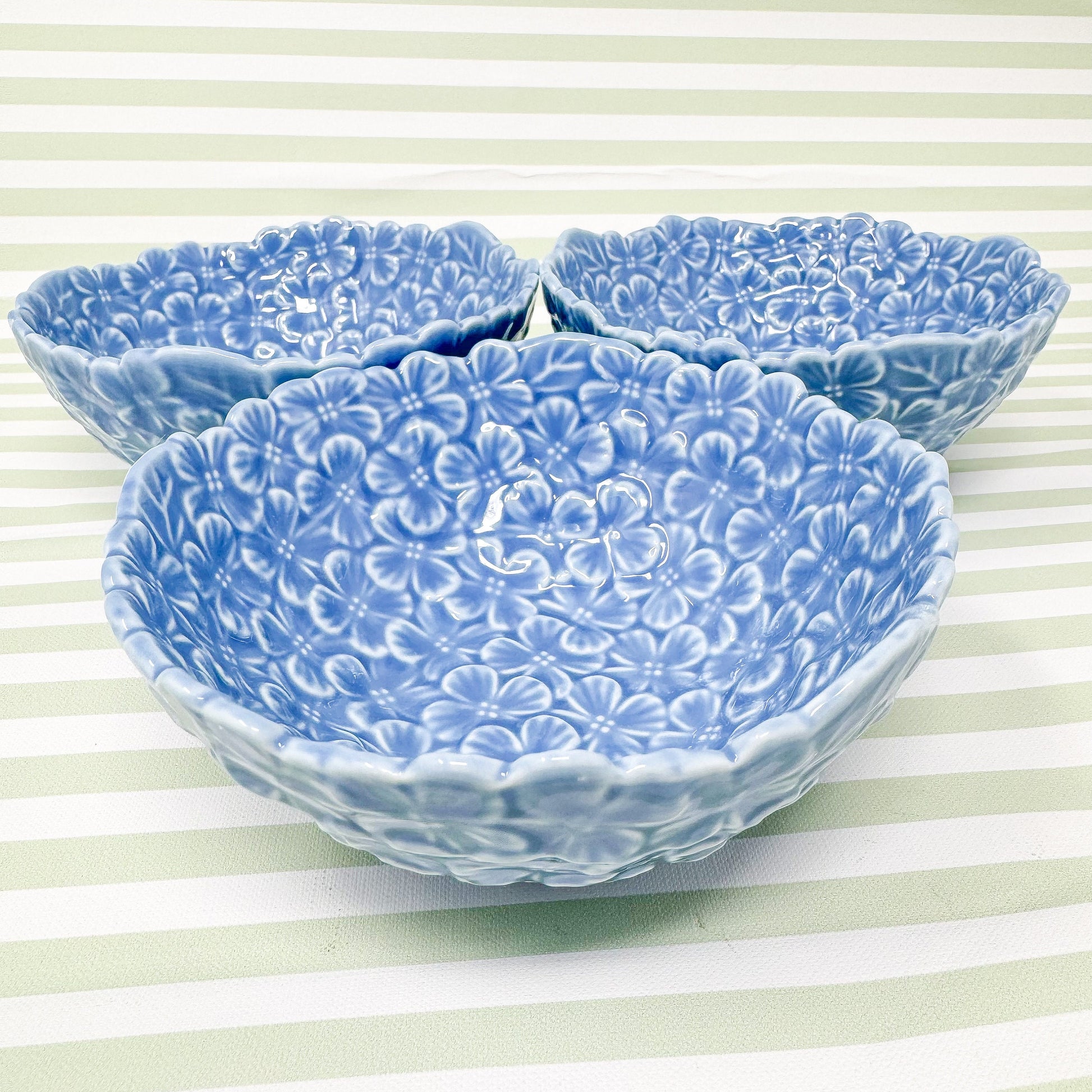 Three light blue ceramic bowls. Each bowl features an embossed floral design of small, light blue flowers covering the entire surface. The bowls are shallow and have a slightly scalloped edge. They appear to be made of a smooth, glazed ceramic.