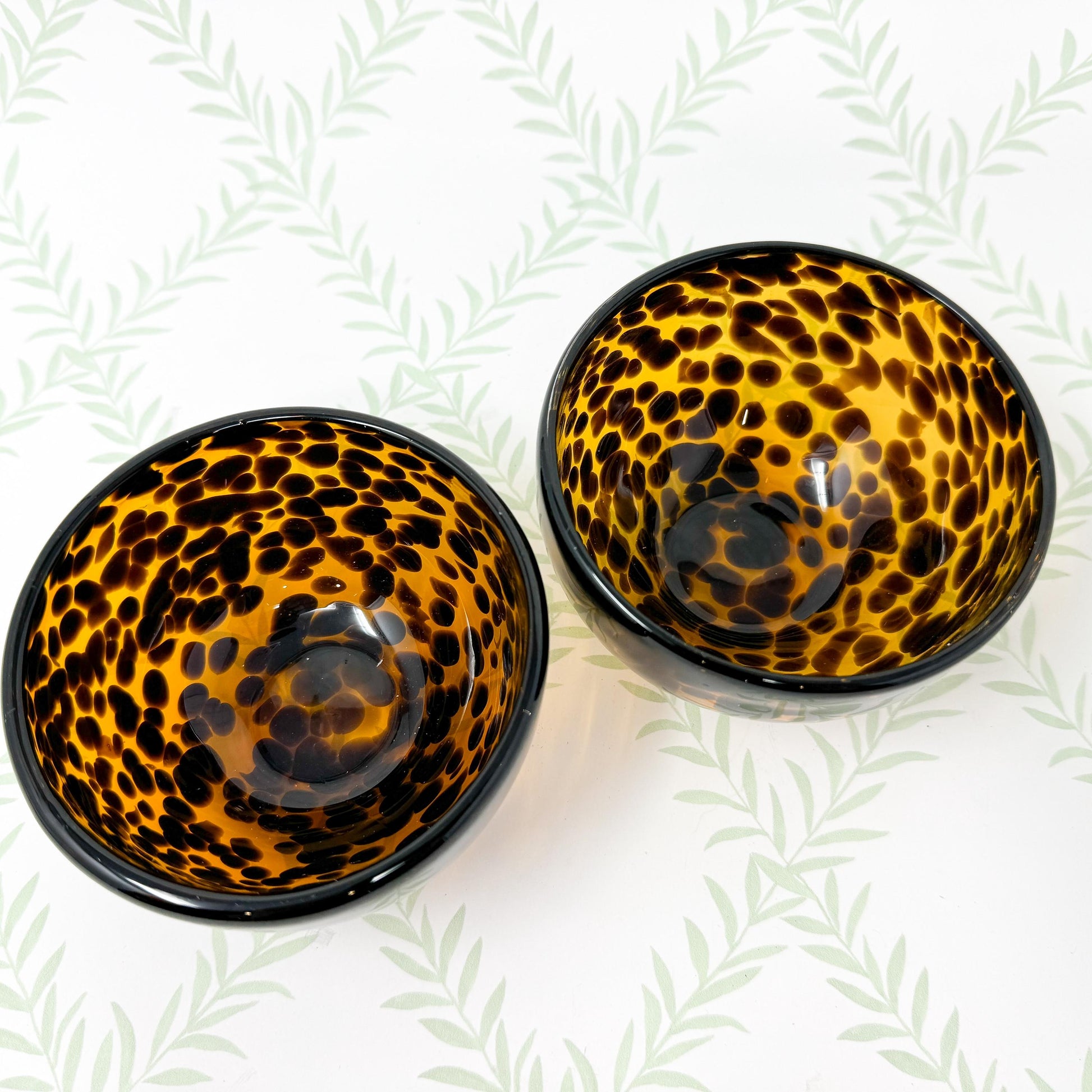 Two small, round bowls with a tortoiseshell pattern. The bowls are made of dark amber glass with dark brown spots, creating a leopard print design.  They are shallow and appear to be decorative or for serving small portions. The bowls are shown against a light green background with a leaf pattern.