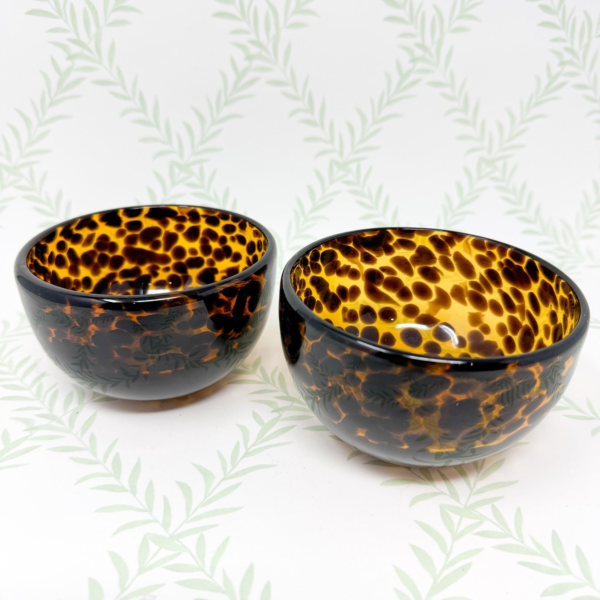 Two small, round bowls with a tortoiseshell pattern. The bowls are made of a dark amber-colored glass or resin with a speckled design resembling a leopard or cheetah print.  They are shallow and have a slightly curved shape. The bowls are displayed against a pale background with a leafy pattern. These decorative bowls are perfect for candy, nuts, or other small items.