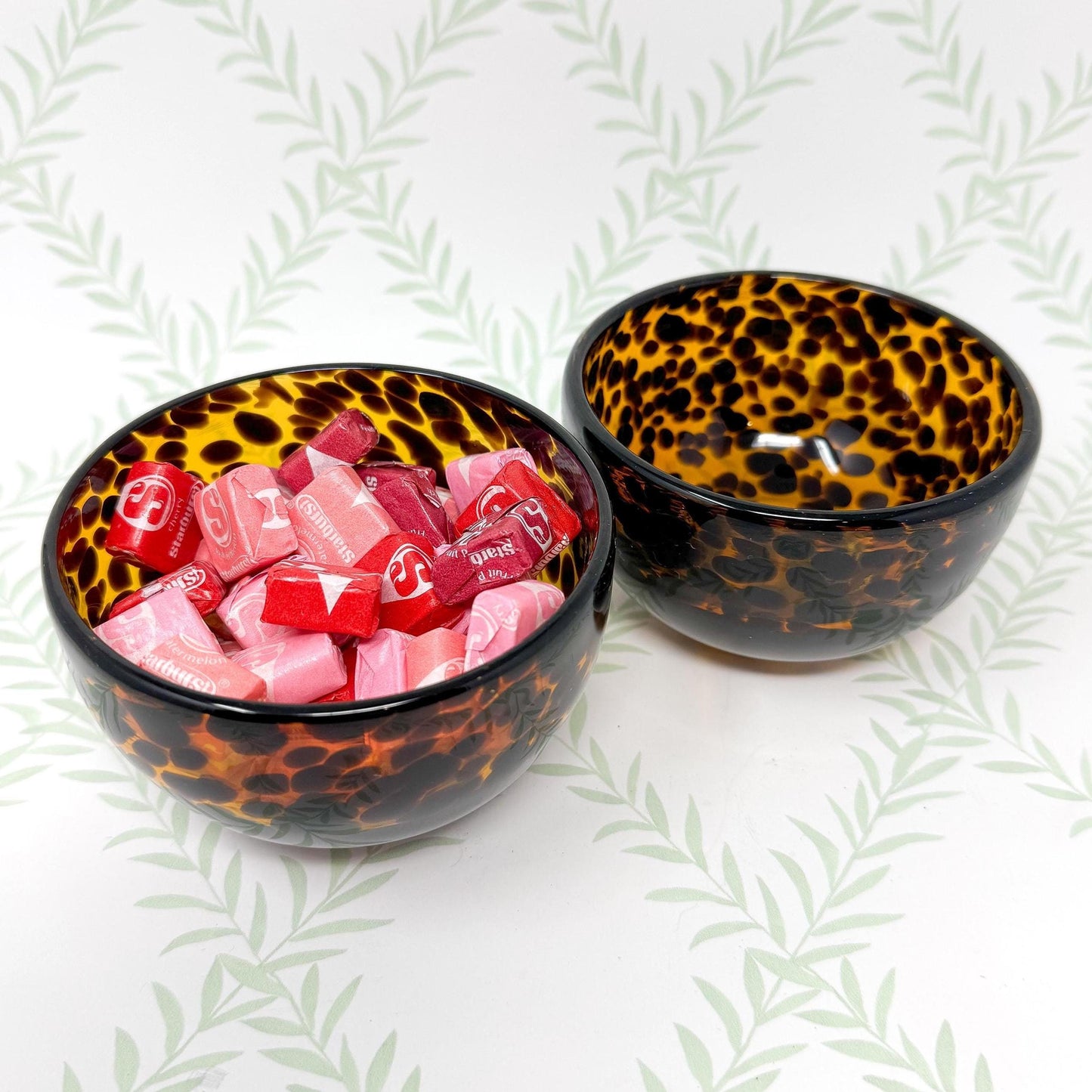 Two small tortoise shell-patterned bowls. One bowl contains several pink and red Starburst candies.  The bowls are glossy and have a dark brown and amber mottled pattern resembling tortoiseshell.  The bowls are shallow and round. The background is a light green with a leafy pattern.